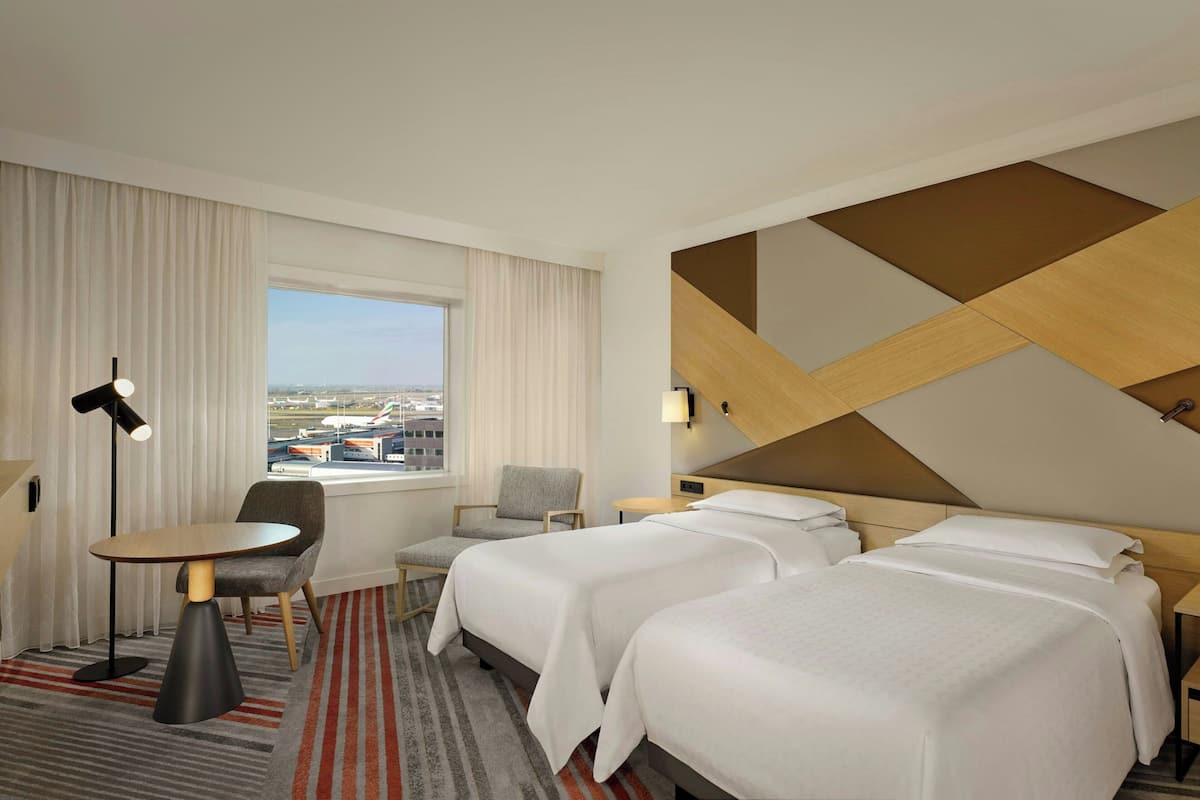 A hotel room with two beds, a desk, a chair, a lamp, and a view of the cityscape.