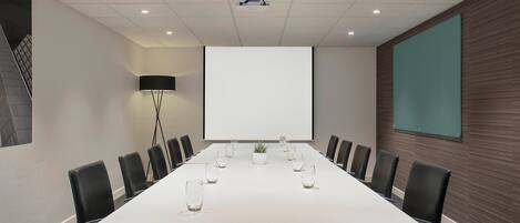 A conference room with a long table, black chairs, a projector screen, and a large screen on the wall.