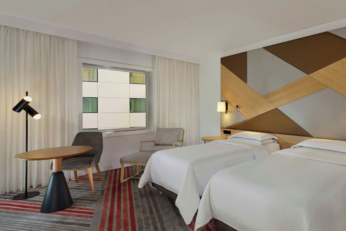 A hotel room with two beds, a dining table, a chair, a lamp, and a window with curtains.