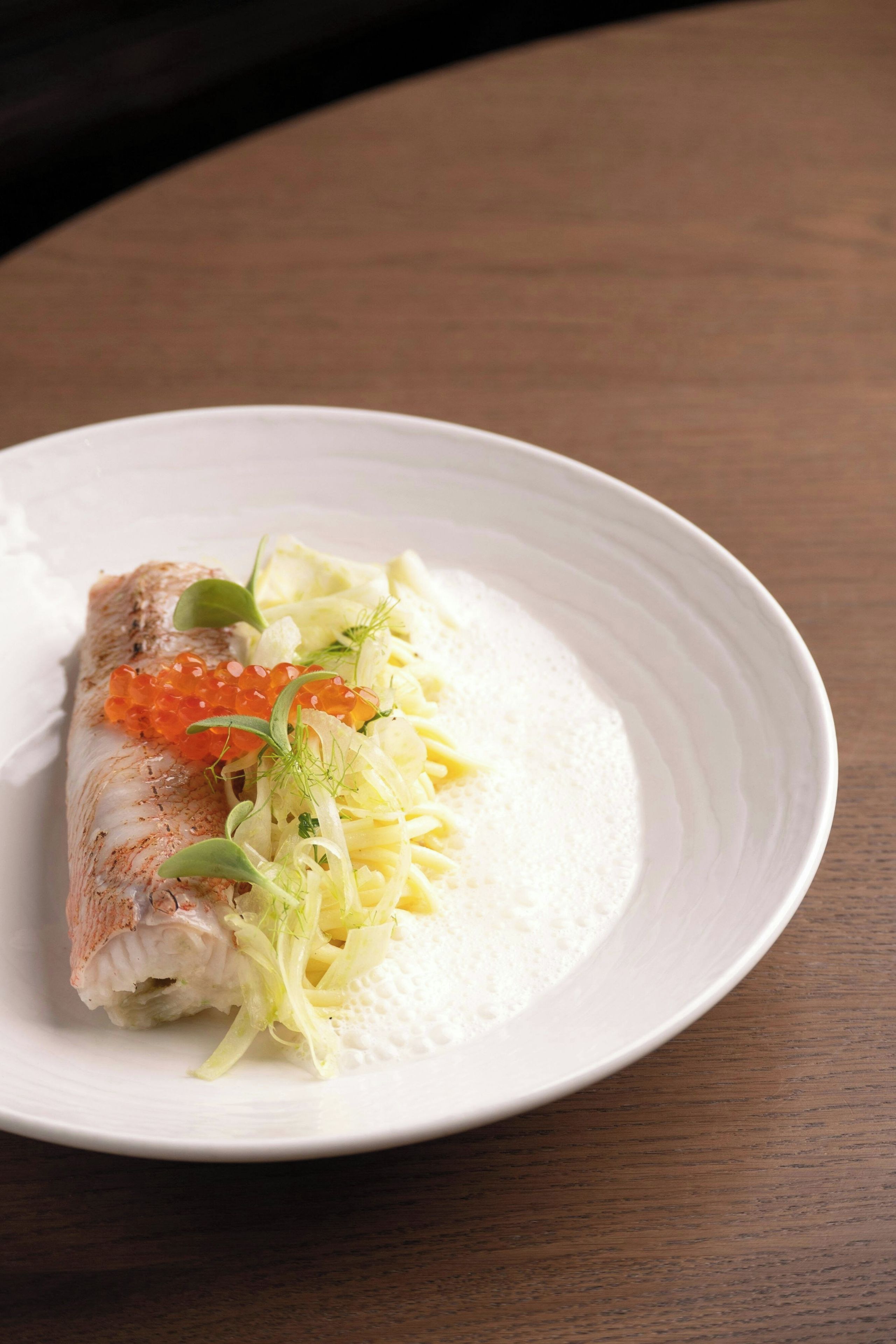 Un plato con un trozo de pescado, adornado con hierbas y huevas, sobre un plato blanco con borde texturizado.