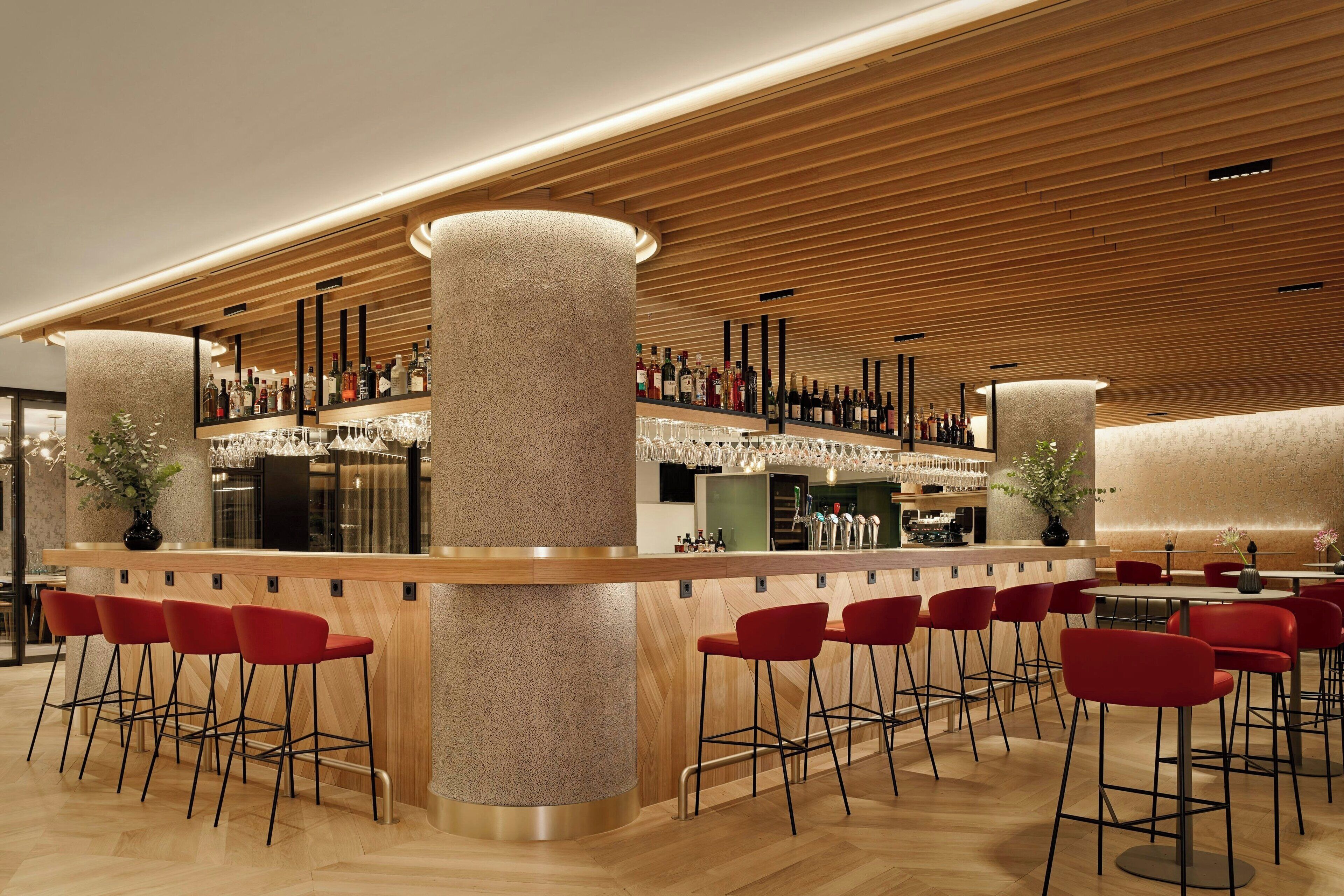 Zona de bar moderna con techo de madera, taburetes rojos y una barra bien surtida.