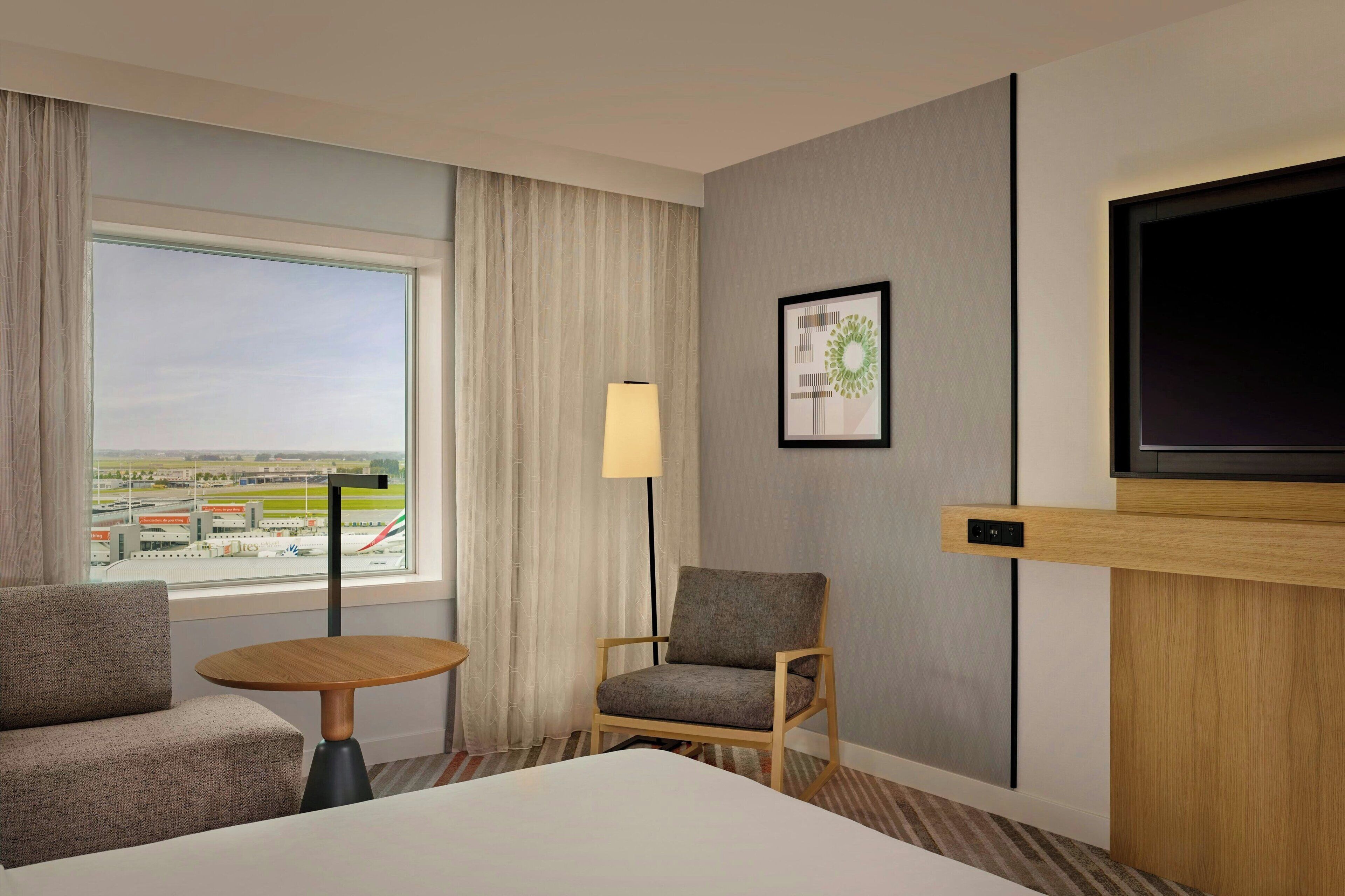 A hotel room with a bed, a sofa, a small table, a chair, a TV, a window with curtains, and a framed picture on the wall.