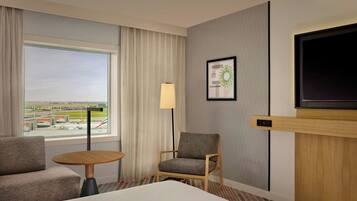 A hotel room with a bed, a sofa, a small table, a chair, a TV, a window with curtains, and a framed picture on the wall.