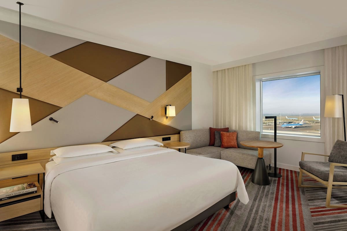 A hotel room with a large bed, a sofa, a small table, and a chair. There is a window with a view of an airport.