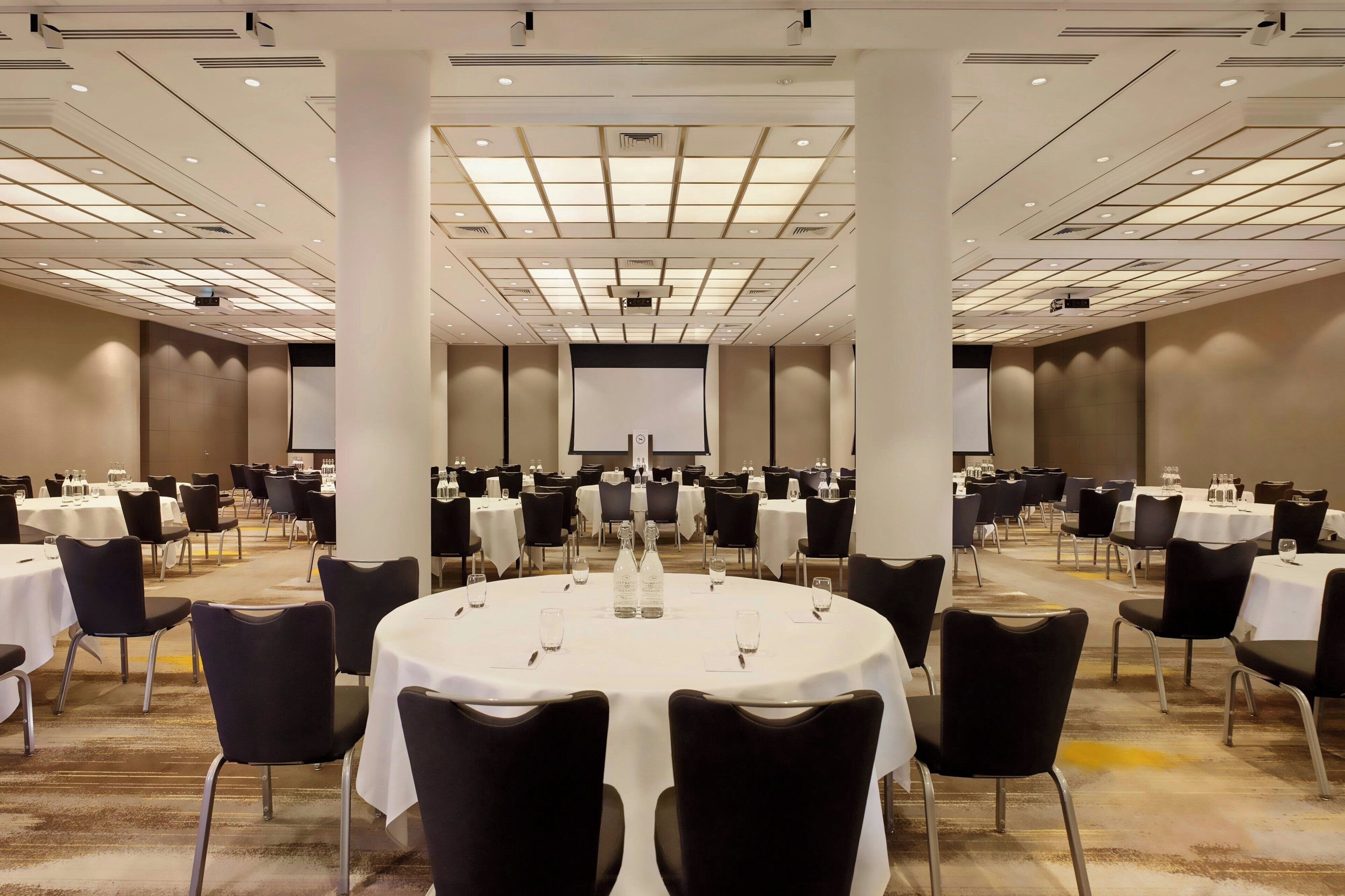 Une grande salle de conférence dotée de tables rondes et de chaises noires, et équipée de bouteilles d’eau et de verres.