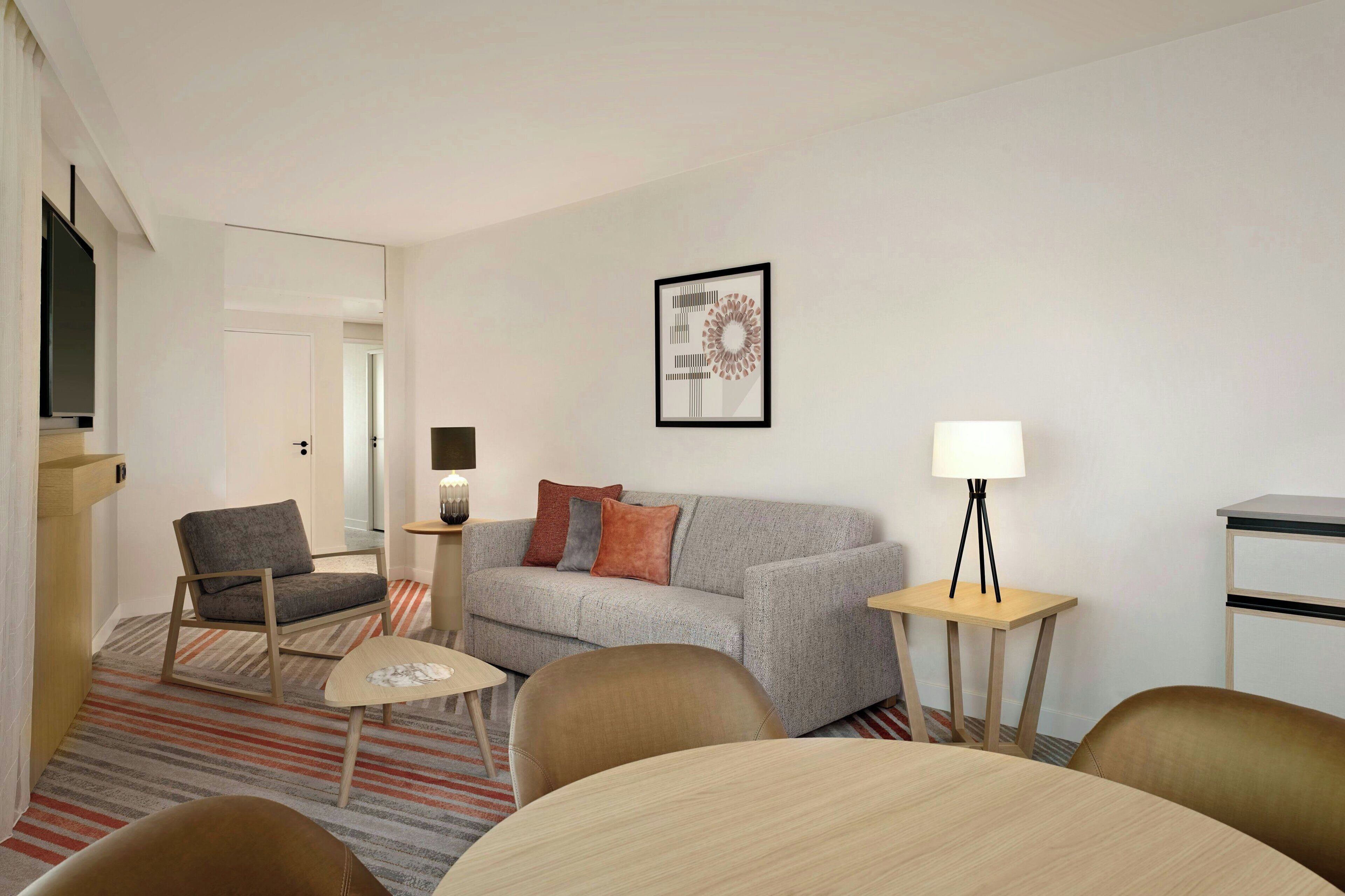 Una habitación de hotel moderna con un sofá gris, una silla de madera, una mesita redonda pequeña y una lámpara en una mesita auxiliar.