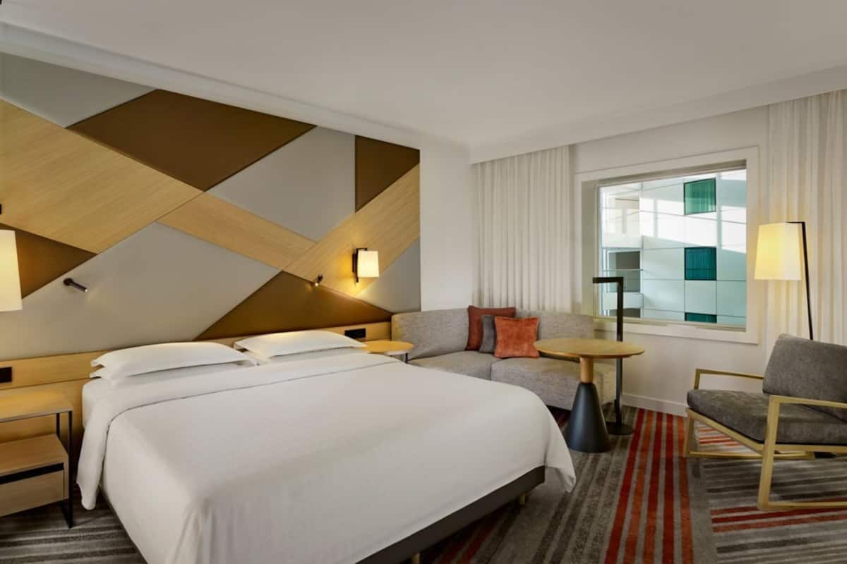A hotel room with a large bed, a sofa, a small table, and a chair. There is a window with curtains and a lamp on the wall.