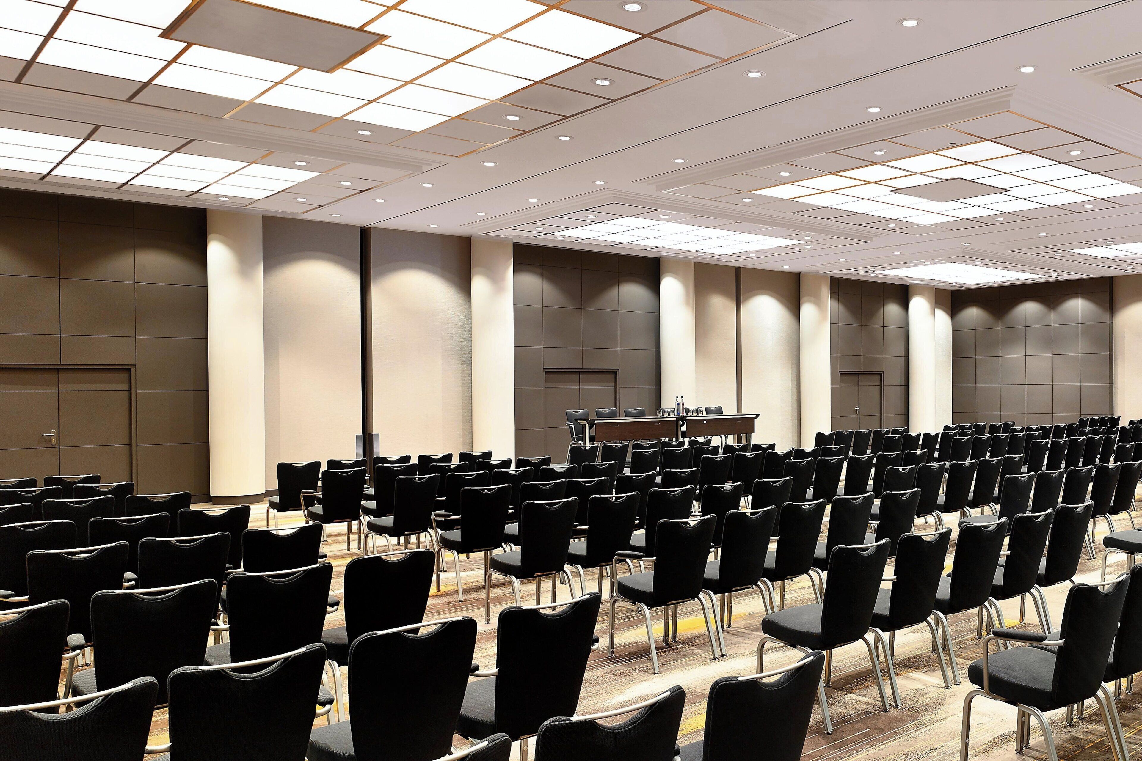 A conference room with rows of black chairs, a podium, and a large ceiling with recessed lighting.