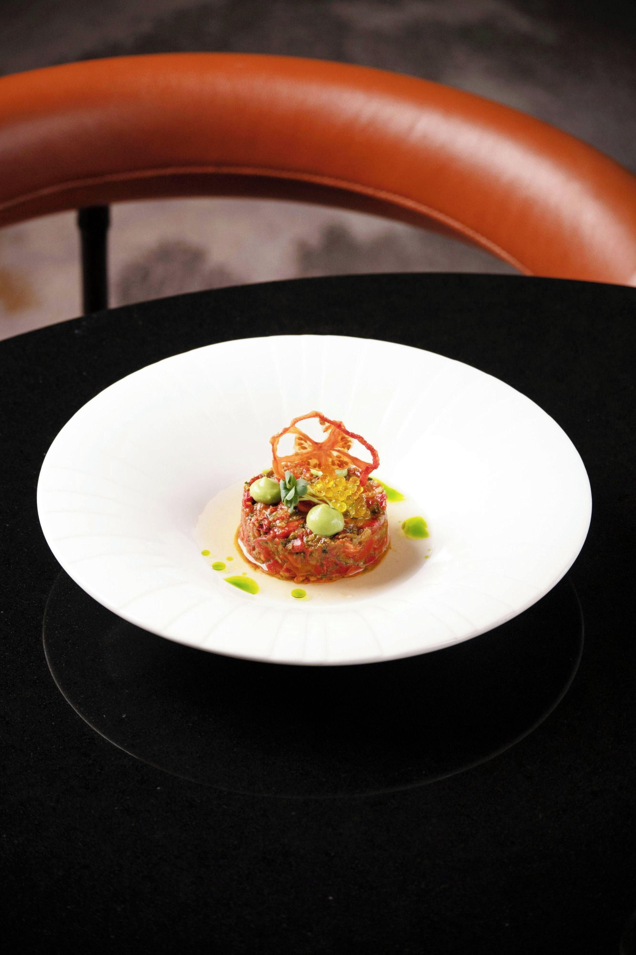 A white plate with a central food item, surrounded by a black table and a brown chair.