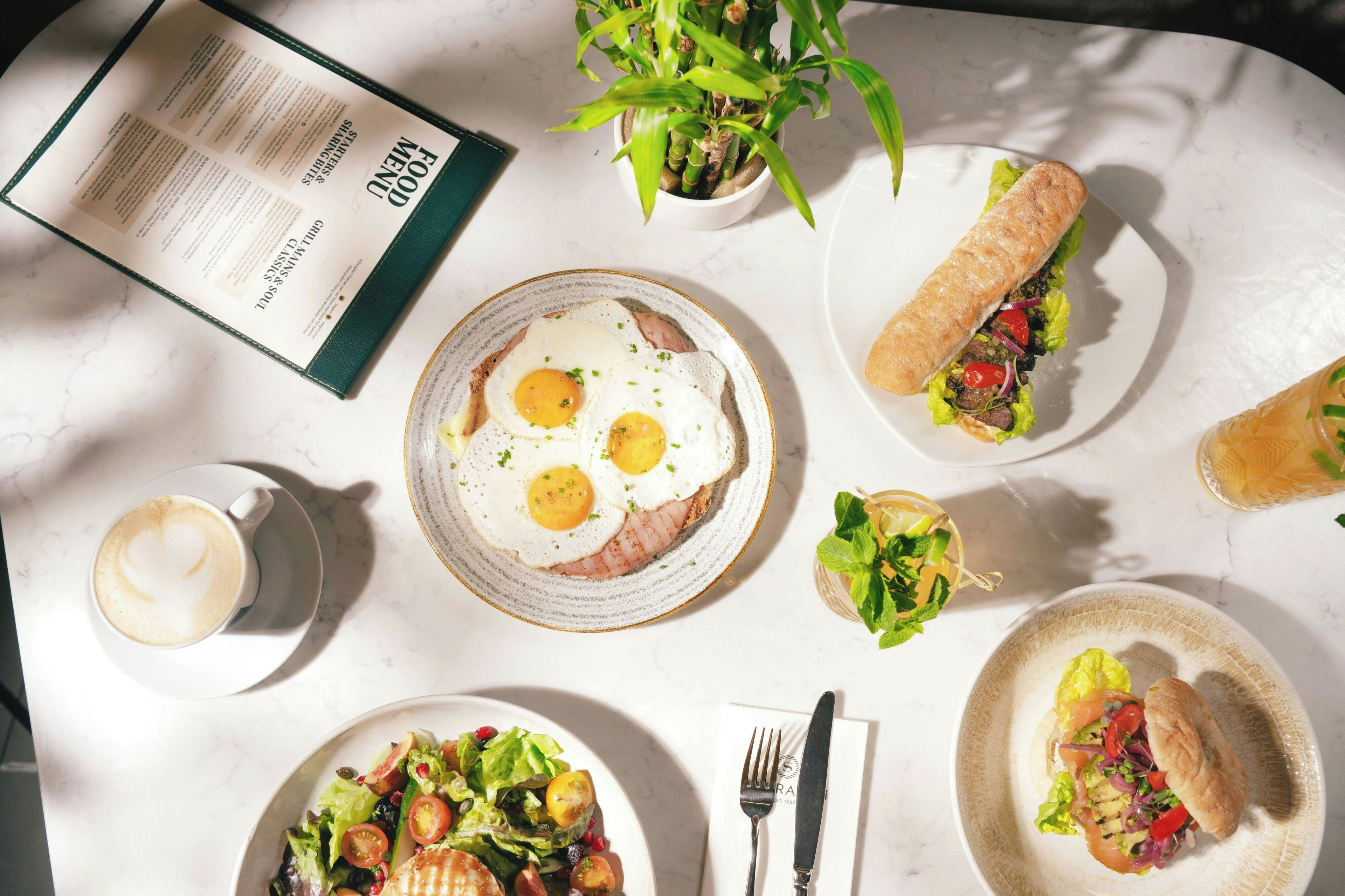 Una mesa con un menú, una taza de café, un plato de huevos, un sándwich, una ensalada y una planta.
