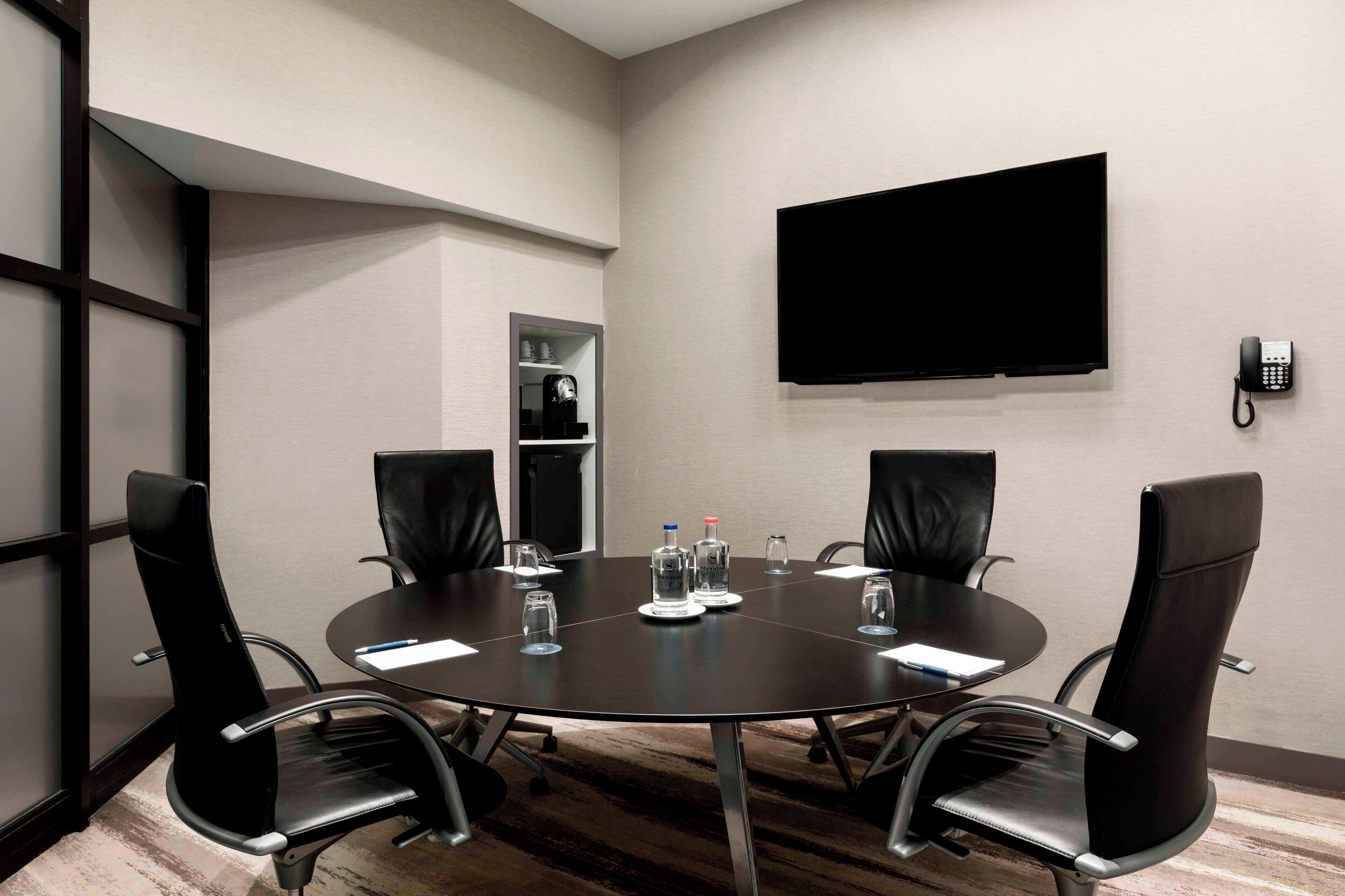 A conference room with a round table, four chairs, a flat-screen TV, a telephone, and a shelf with a coffee machine and water bottles.