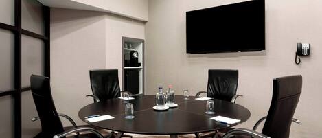 A conference room with a round table, four chairs, a flat-screen TV, a telephone, and a shelf with a coffee machine and water bottles.