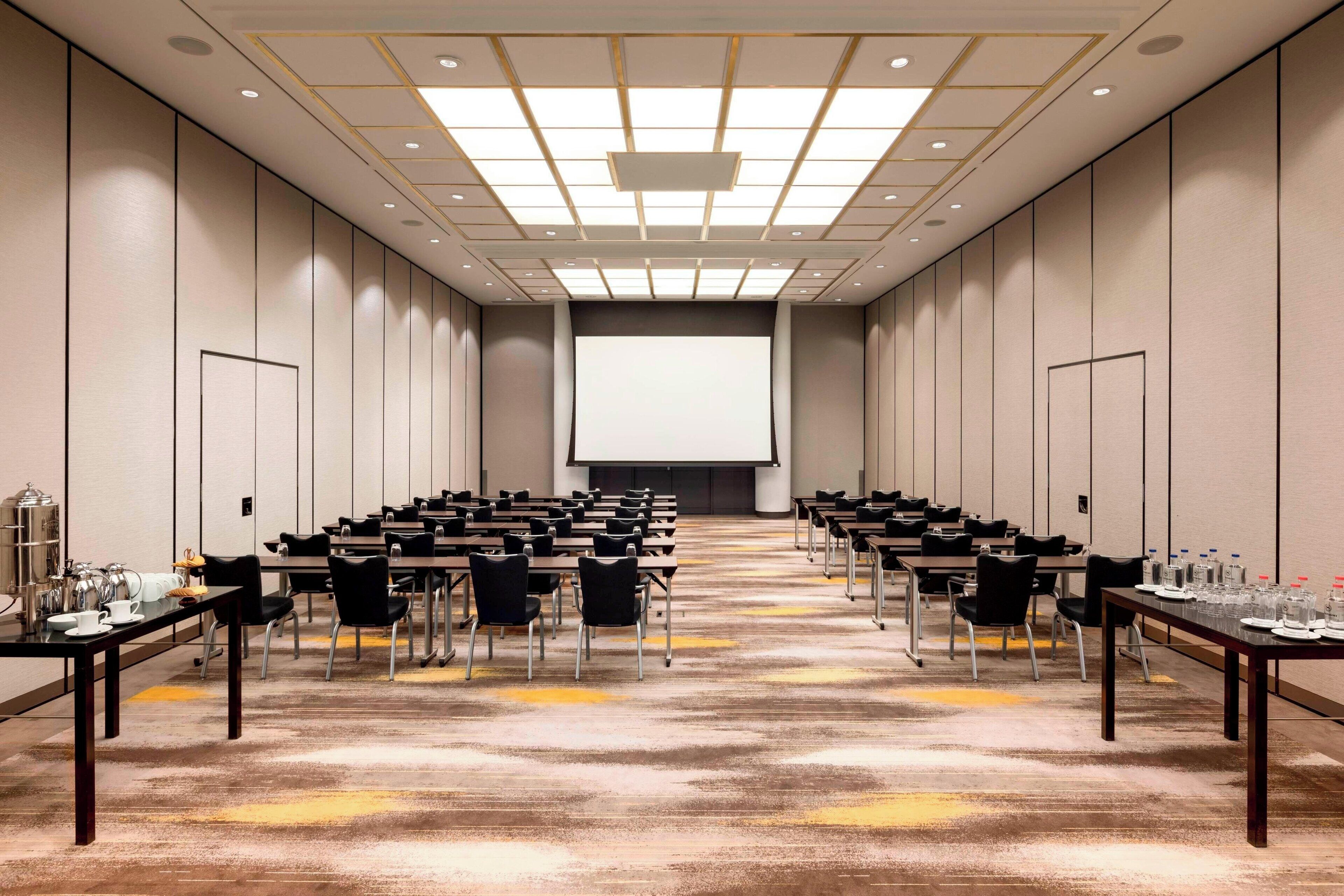 A conference room with rows of tables and chairs, a large screen at the front, and a carpeted floor.