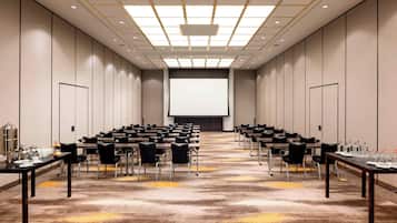 A conference room with rows of tables and chairs, a large screen at the front, and a carpeted floor.