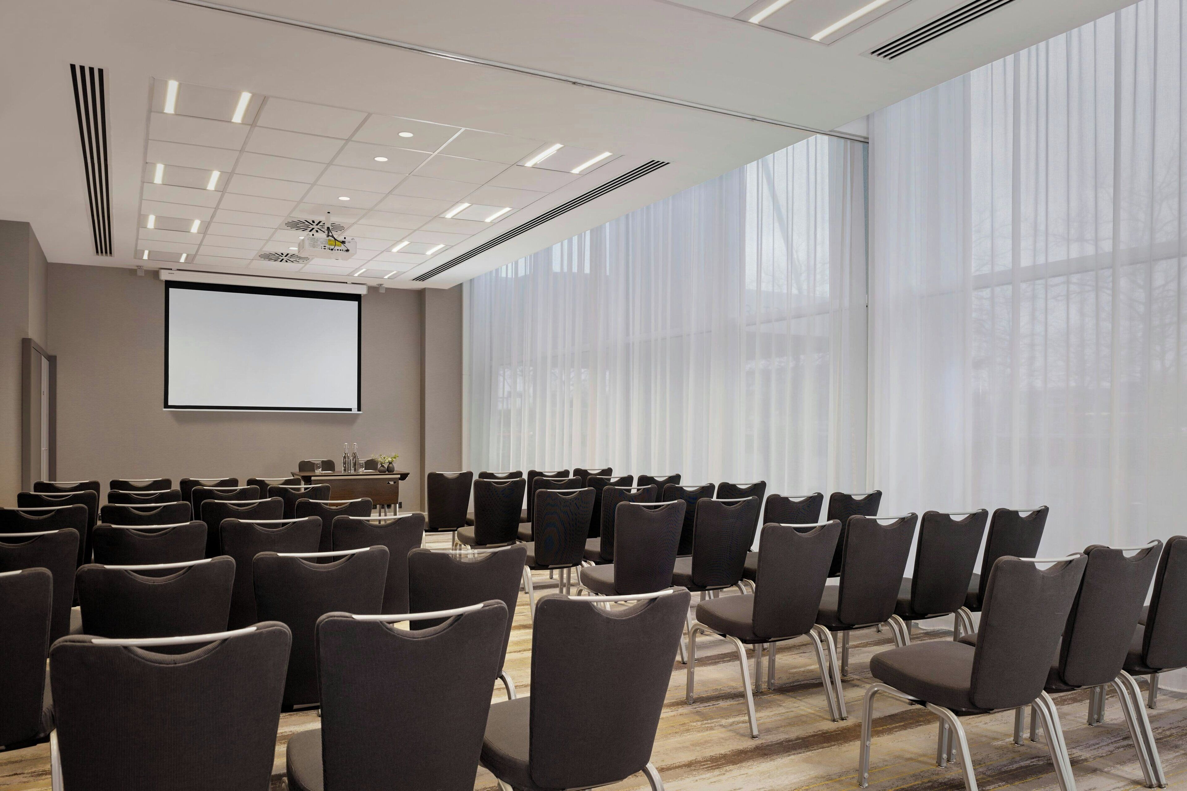 Uma sala de conferência com fileiras de cadeiras, uma tela de projetor e amplas janelas com cortinas leves.