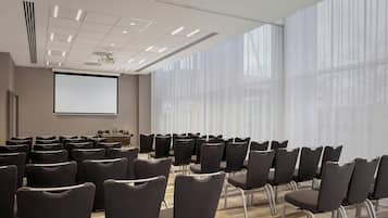 Uma sala de conferência com fileiras de cadeiras, uma tela de projetor e amplas janelas com cortinas leves.