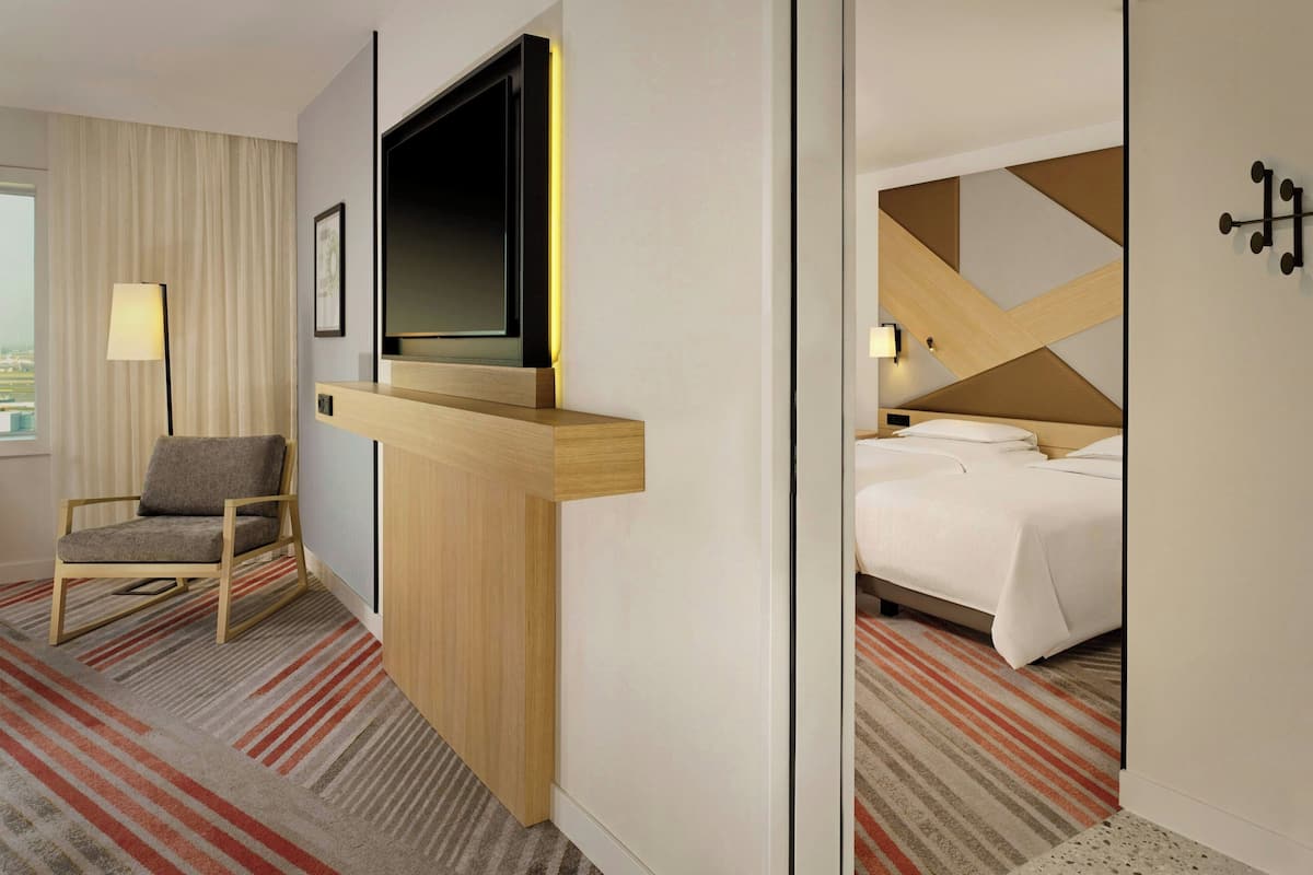 A hotel room with a bed, a TV mounted on a wooden panel, a chair, and a view of the city through the window.