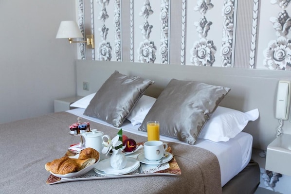 Una cama bien hecha con almohadas grises, una mesita de noche con una lámpara y una bandeja con artículos de desayuno, incluyendo croissants, fruta y un vaso de zumo de naranja.