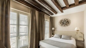 A hotel room with a large bed, a nightstand, a lamp, and a view of the city through large windows. - Hotel la Residence du Berry (Versailles)