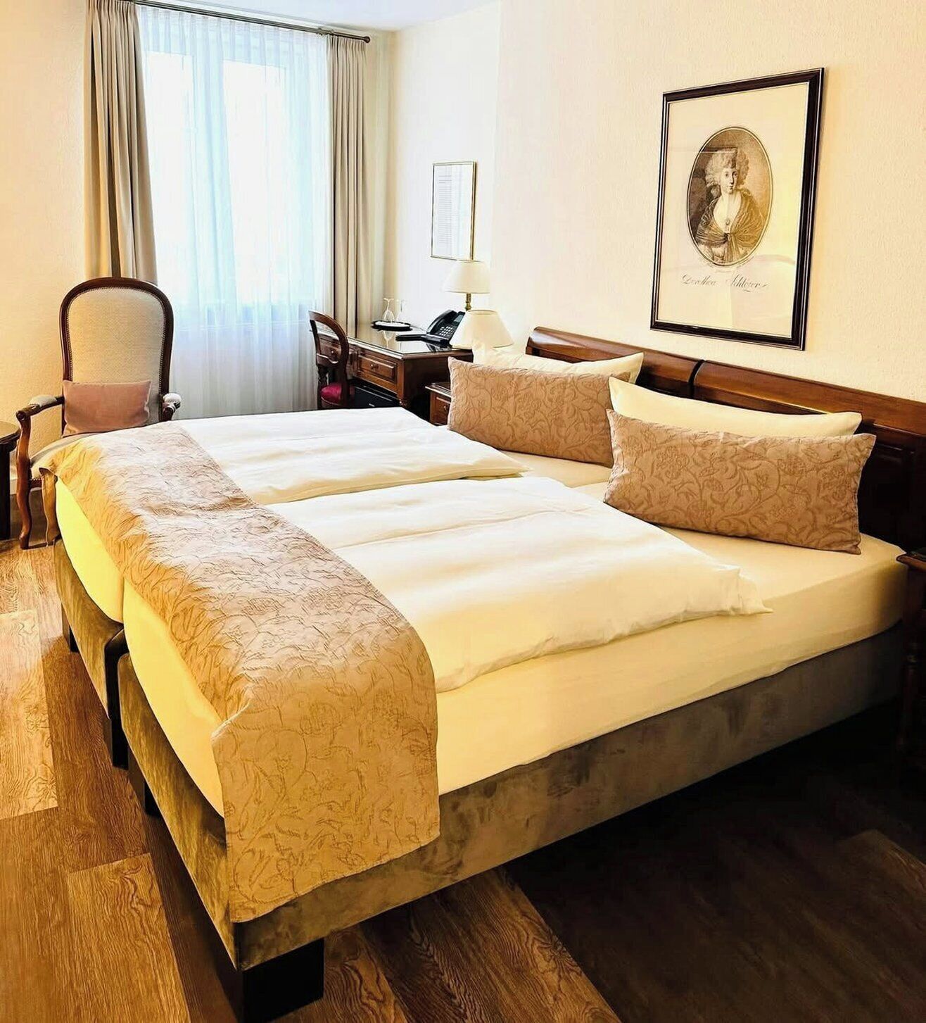 A double bed with a wooden frame, two bedside tables, a chair, a desk, and a framed portrait on the wall.