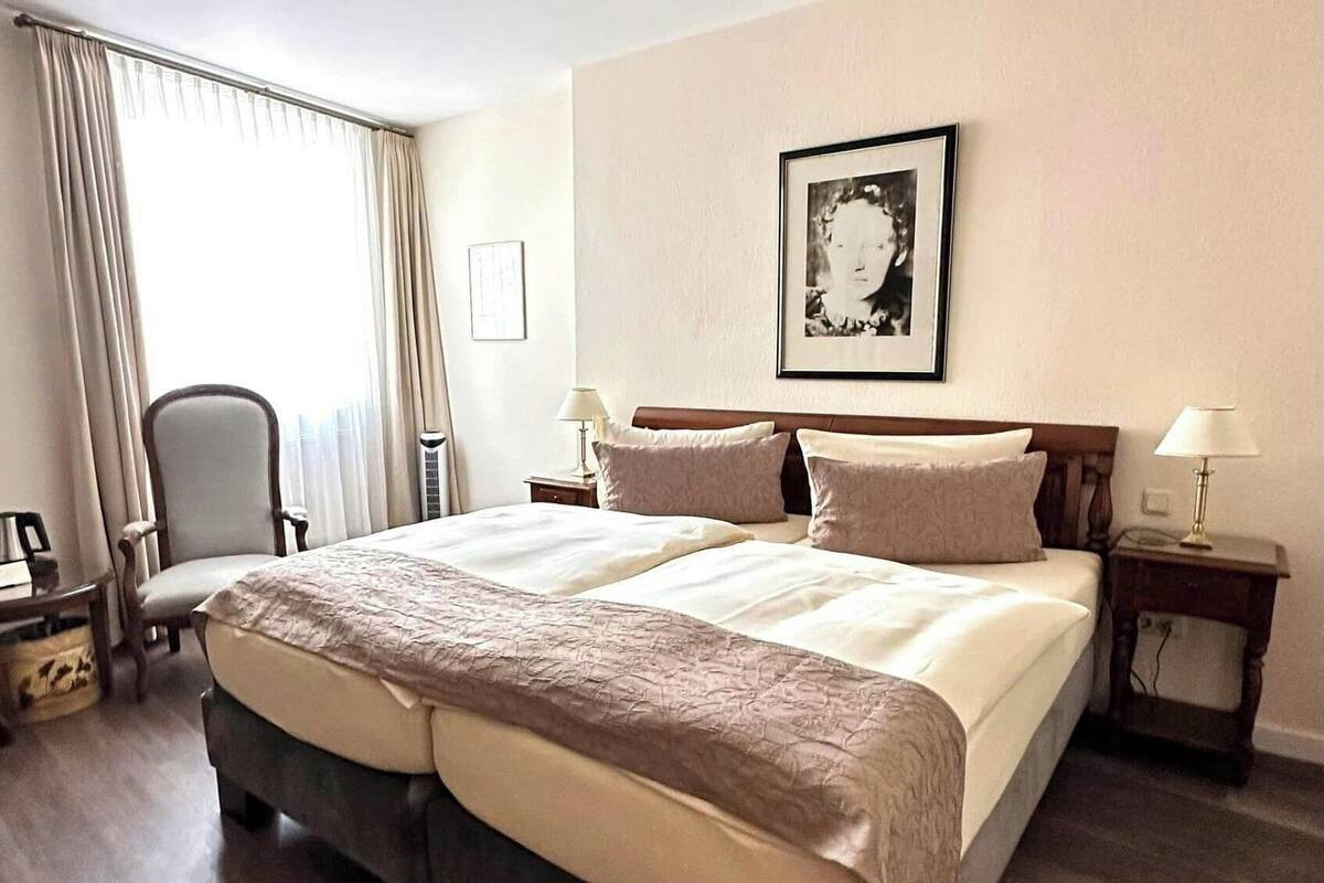 Uma cama de casal com cabeceira escura e duas mesas de cabeceira, cada uma com uma lâmpada. Uma cadeira está posicionada aos pés da cama, e um quadro emoldurado está pendurado na parede.