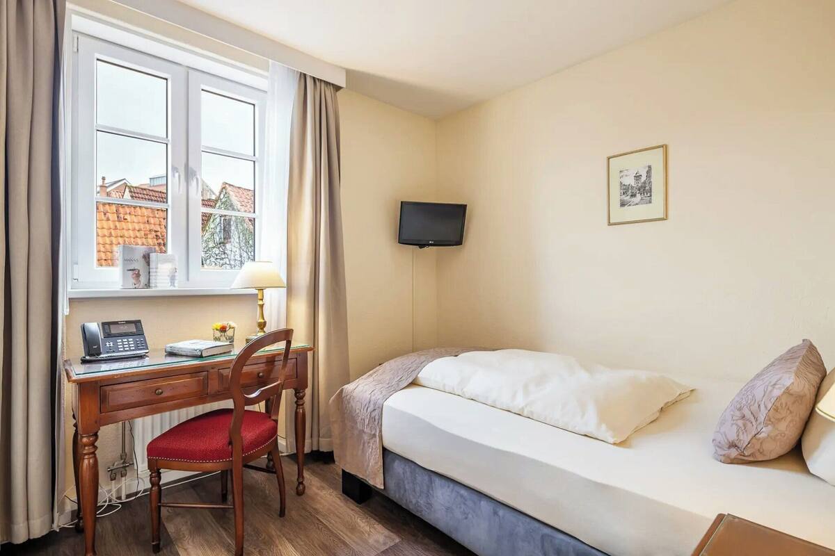 Quarto de hotel com cama, escrivaninha com telefone, cadeira, televisão fixada na parede e janela com cortinas.