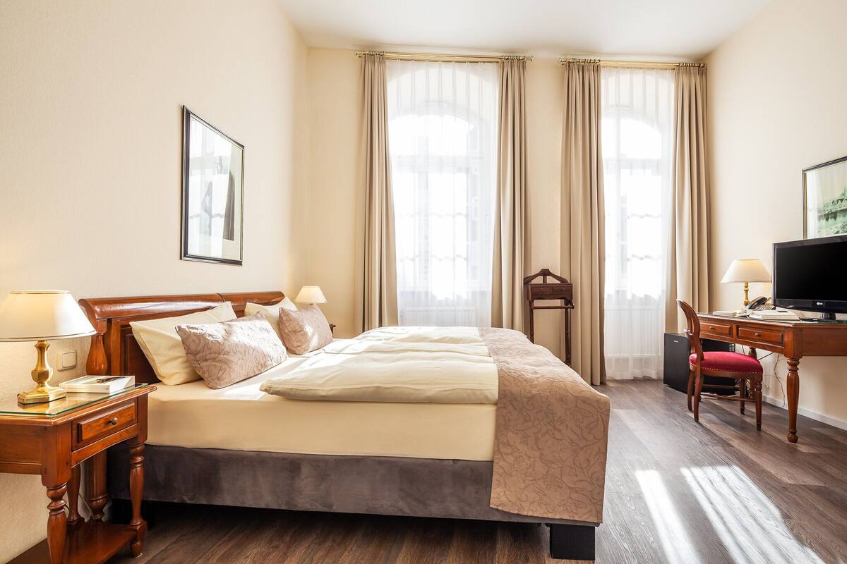 Quarto de hotel com cama, mesas de cabeceira, escrivaninha, cadeira, televisão e janelas amplas com cortinas.