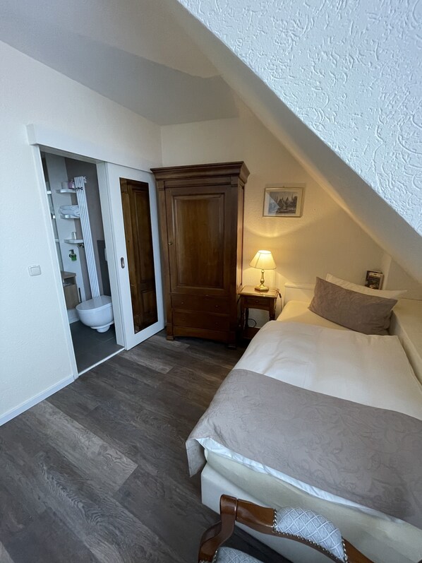 Un dormitorio con cama, armario de madera, mesita de noche con lámpara y baño visible a través de una puerta abierta.