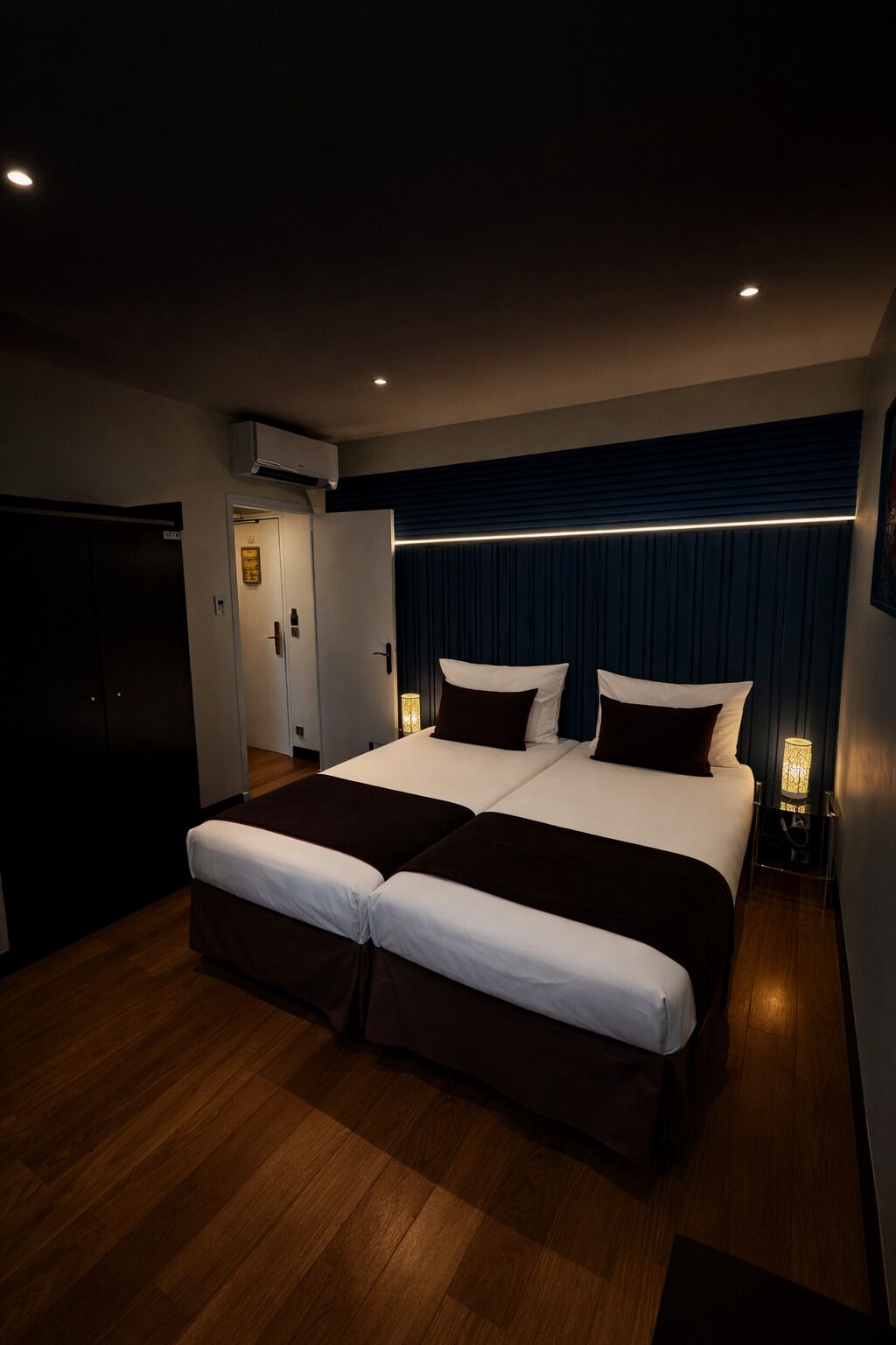 Una cama doble con cabecera oscura, una mesita de noche con lámpara y suelo de madera.
