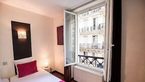 A hotel room with a bed, a nightstand, a wall-mounted lamp, a window with a view of a building, and a radiator. - Hotel Pax Opera (Paris)