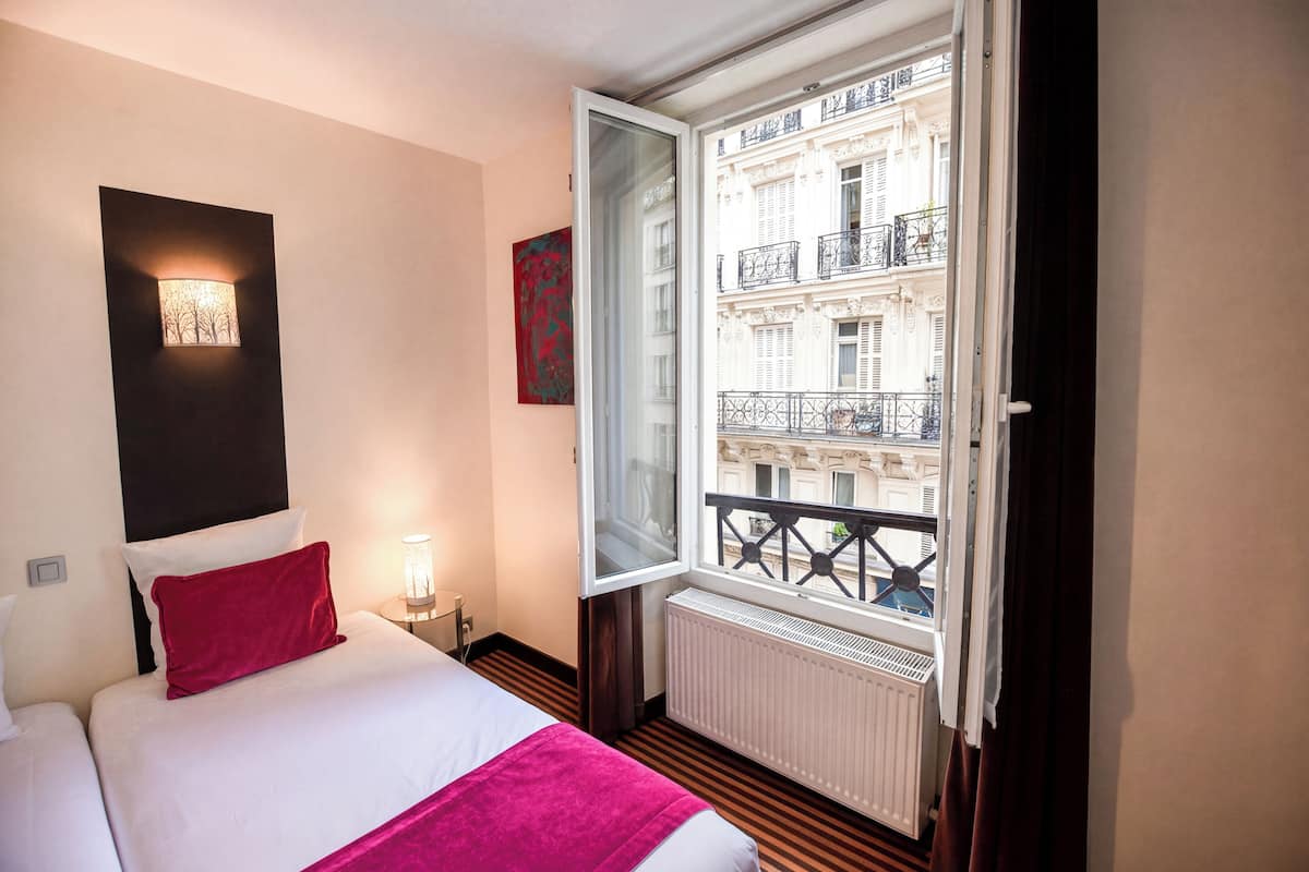 Habitación de hotel con cama, mesita de noche, lámpara de pared, ventana con vista a un edificio y radiador.