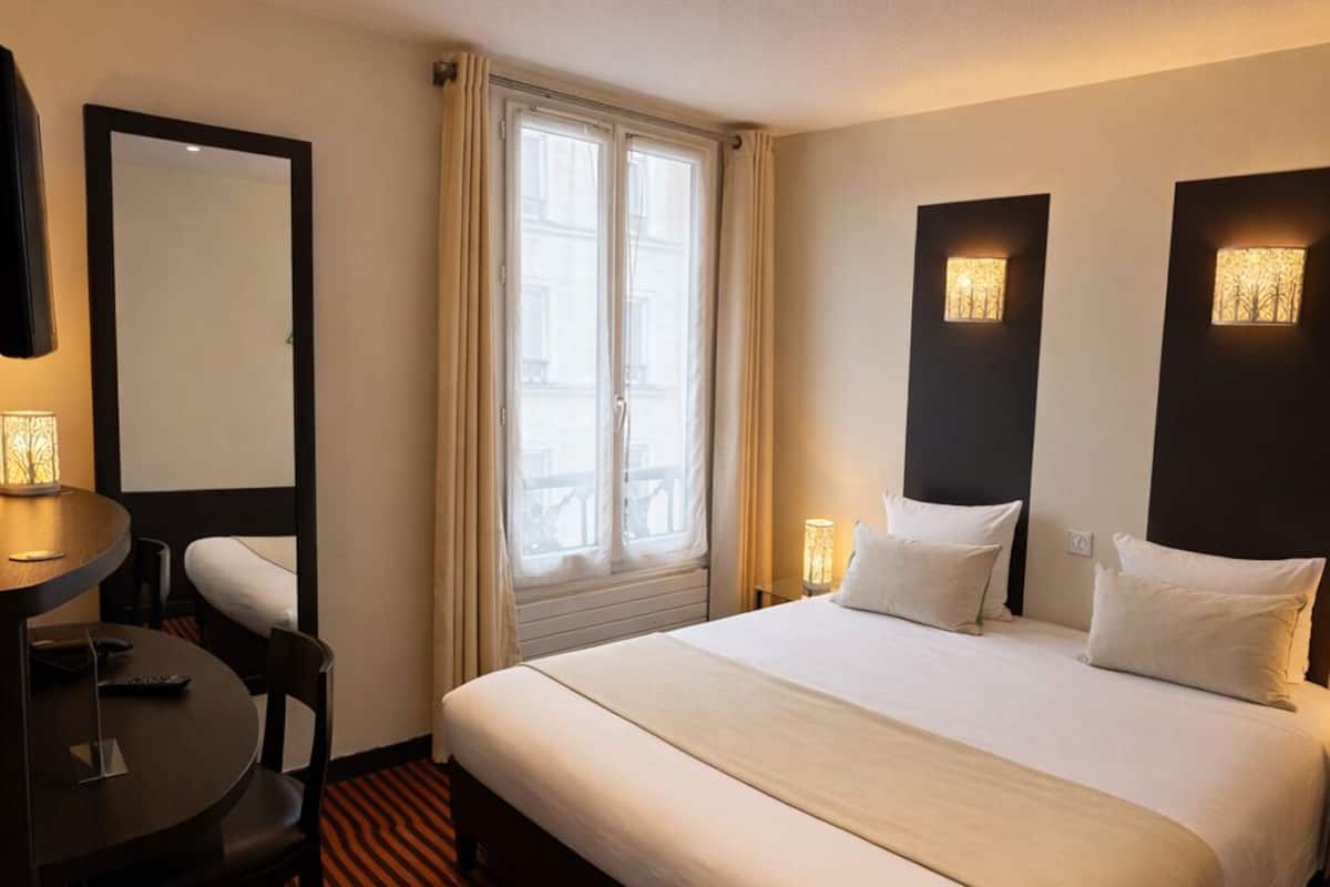 Una habitación de hotel con una cama grande, una mesa redonda con una silla, un espejo y dos lámparas de pared.