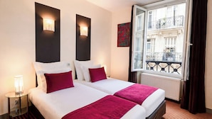 A hotel room with two beds, a nightstand, a lamp, a window with a view of buildings, and a radiator. - Hotel Pax Opera (Paris)