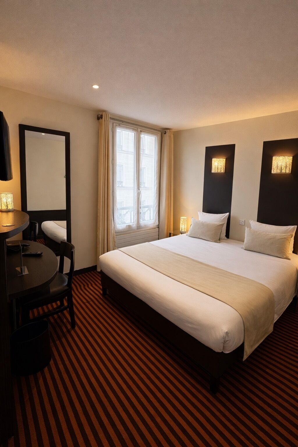 Une chambre d’hôtel avec un grand lit, une table ronde avec une chaise, un miroir et deux lampes fixées au mur.