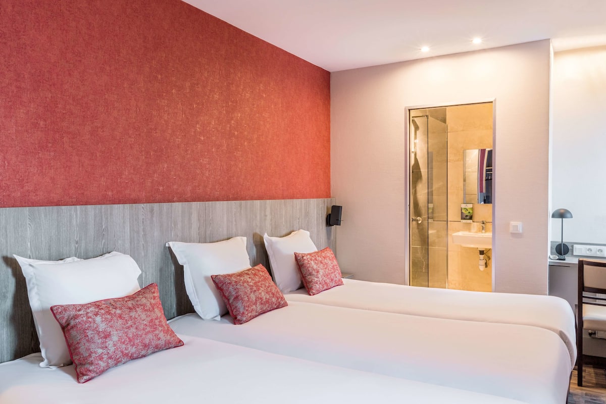 A hotel room with two beds, a red accent wall, a wooden headboard, and a glass shower enclosure.