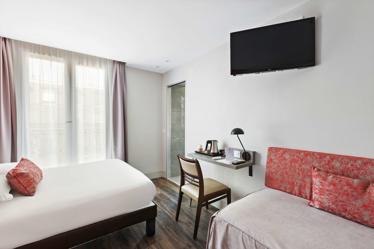 A hotel room with a bed, a desk, a chair, a sofa, a TV, and a window with curtains.