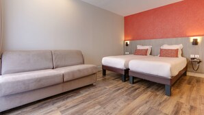 Una habitación de hotel moderna con un sofá color beige, dos camas individuales, suelo de madera y una pared de acento roja.
