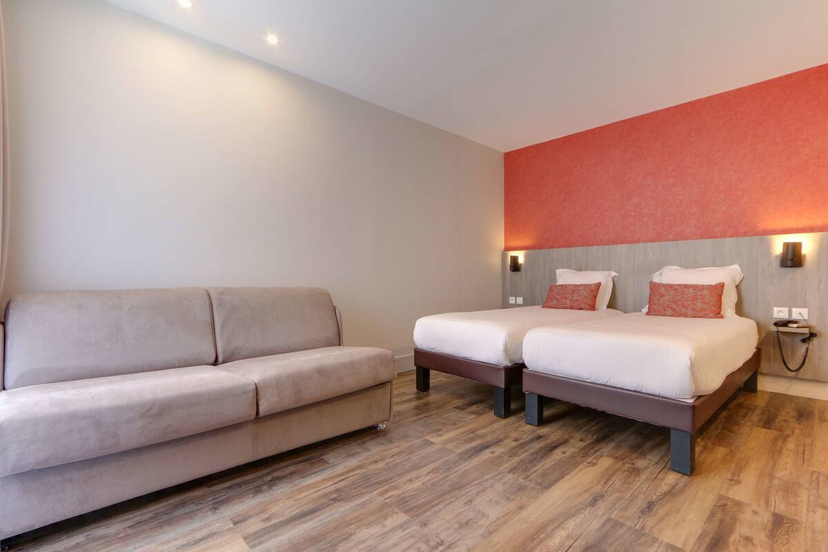 A modern hotel room with a beige sofa, two single beds, wooden flooring, and a red accent wall.