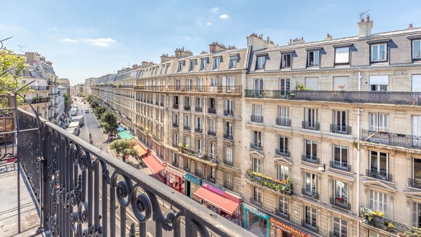 Una hilera de edificios de estilo europeo con balcones, una calle con tiendas y un cielo azul despejado.
