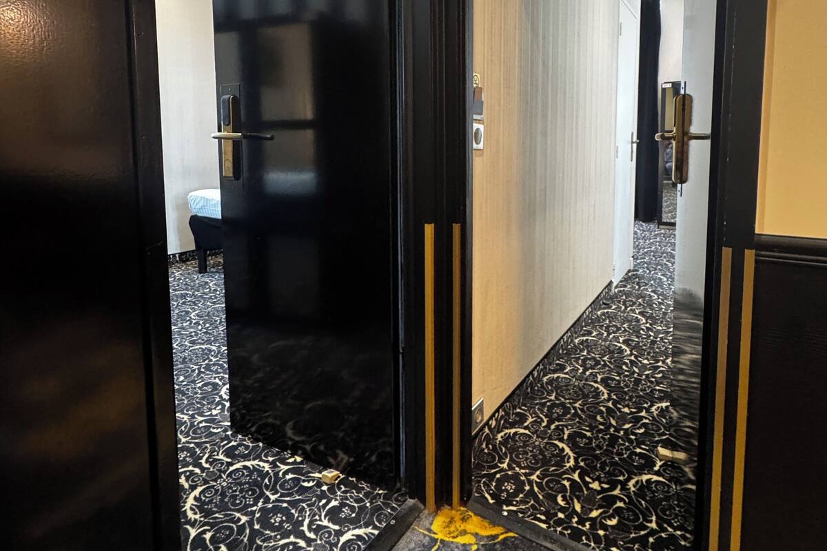A hotel room with a black and white patterned carpet, a black door with a yellow sign, and a glimpse of a bed and a bathroom in the background.