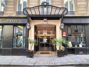 The entrance of a hotel with a black facade, a sign reading "Opéra", and two potted plants at the entrance. - Hotel Peyris Opera (Paris)