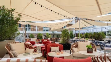 Relax in the shaded outdoor seating area with comfortable chairs and tables, surrounded by lush greenery and modern architecture.