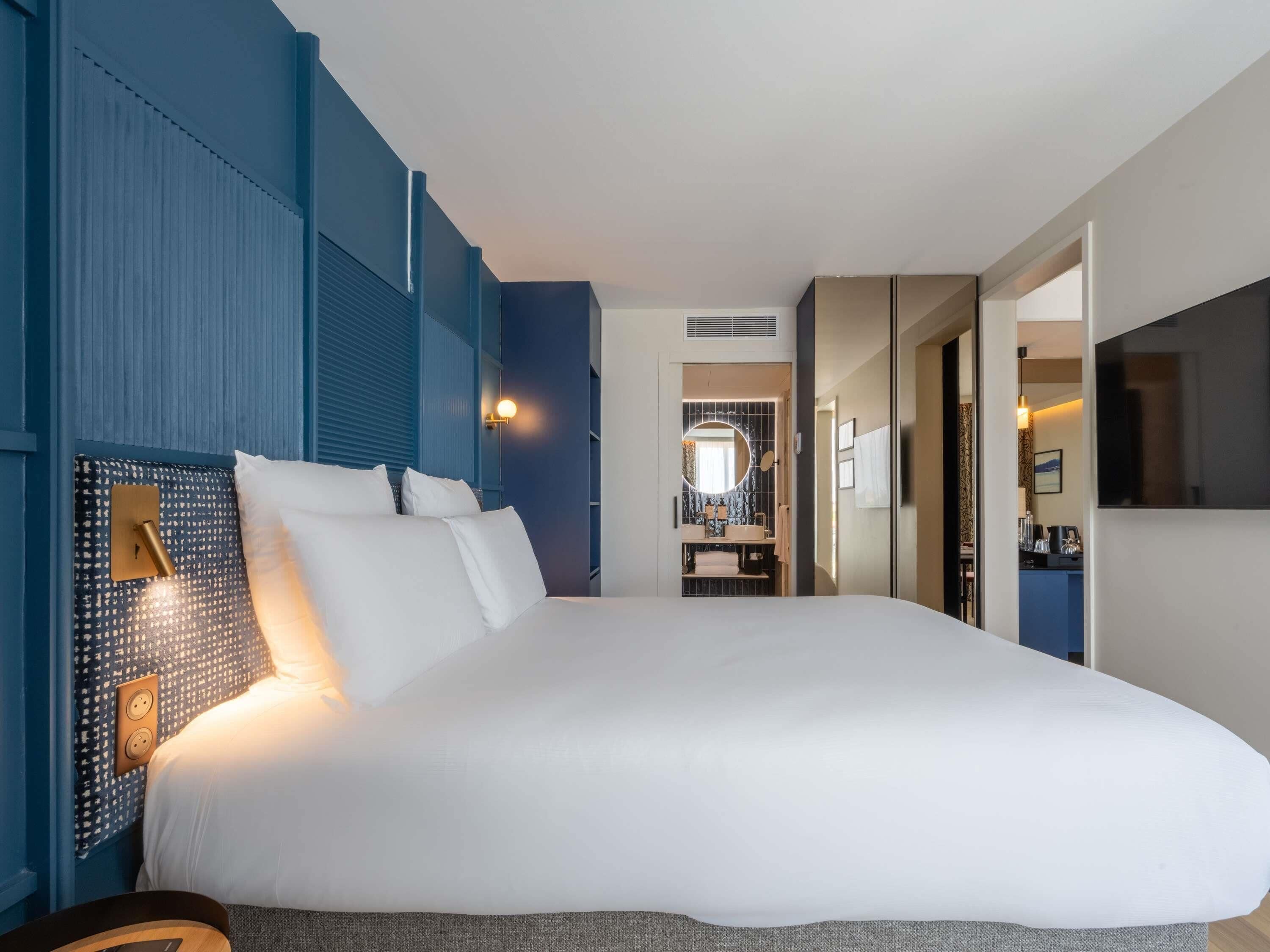 Habitación de hotel con una cama grande, una lámpara de noche, un espejo y vistas al baño.