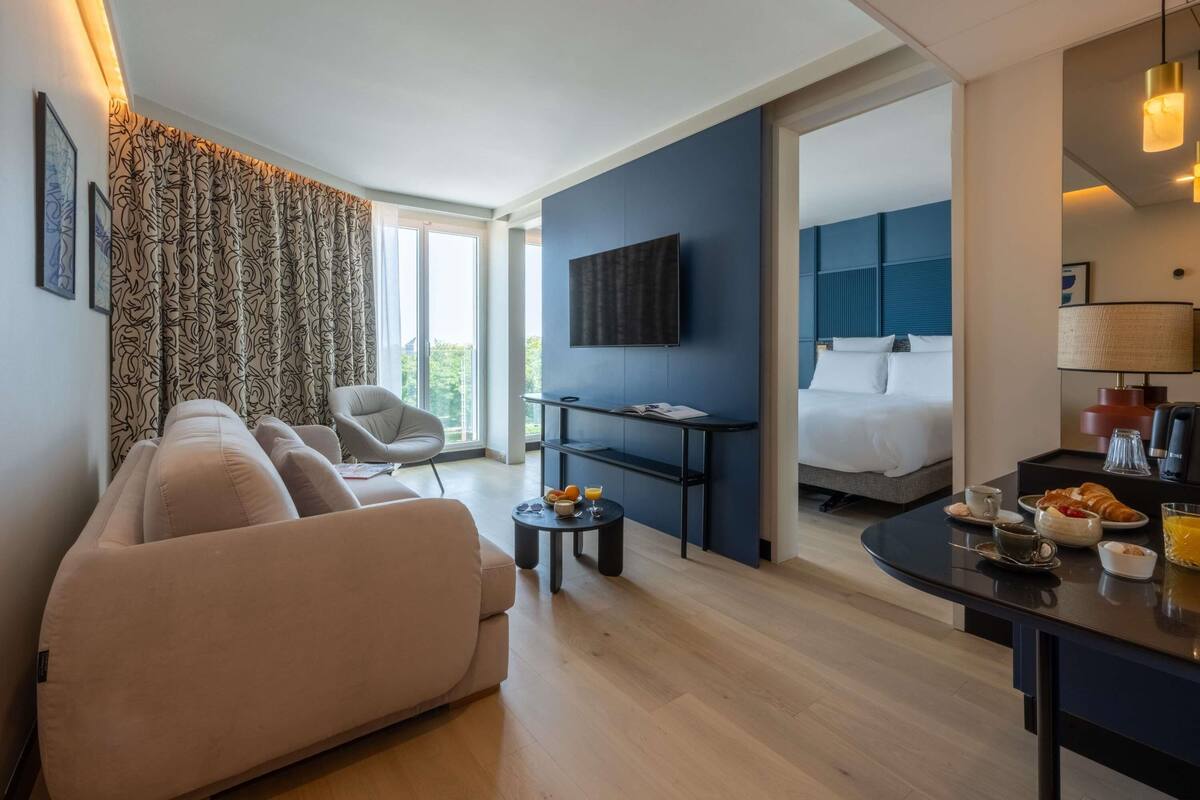 Une chambre d’hôtel moderne avec un canapé, une table à manger avec des articles pour le petit-déjeuner, une télévision et un lit avec une tête de lit bleue.