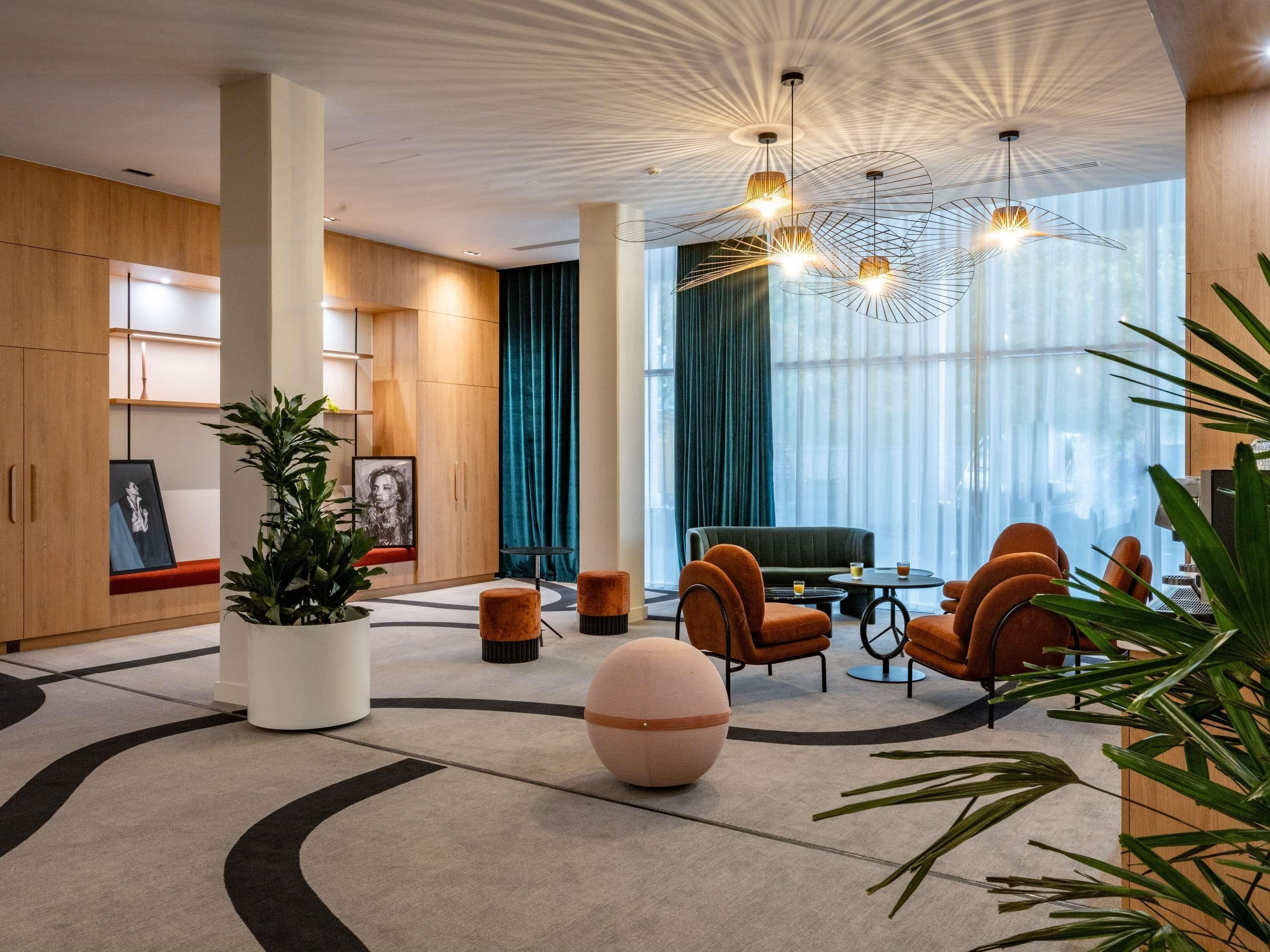 Un hall d’hôtel moderne avec des panneaux de bois, une grande plante en pot, un pouf rond, des fauteuils orange et un luminaire au plafond au design original.