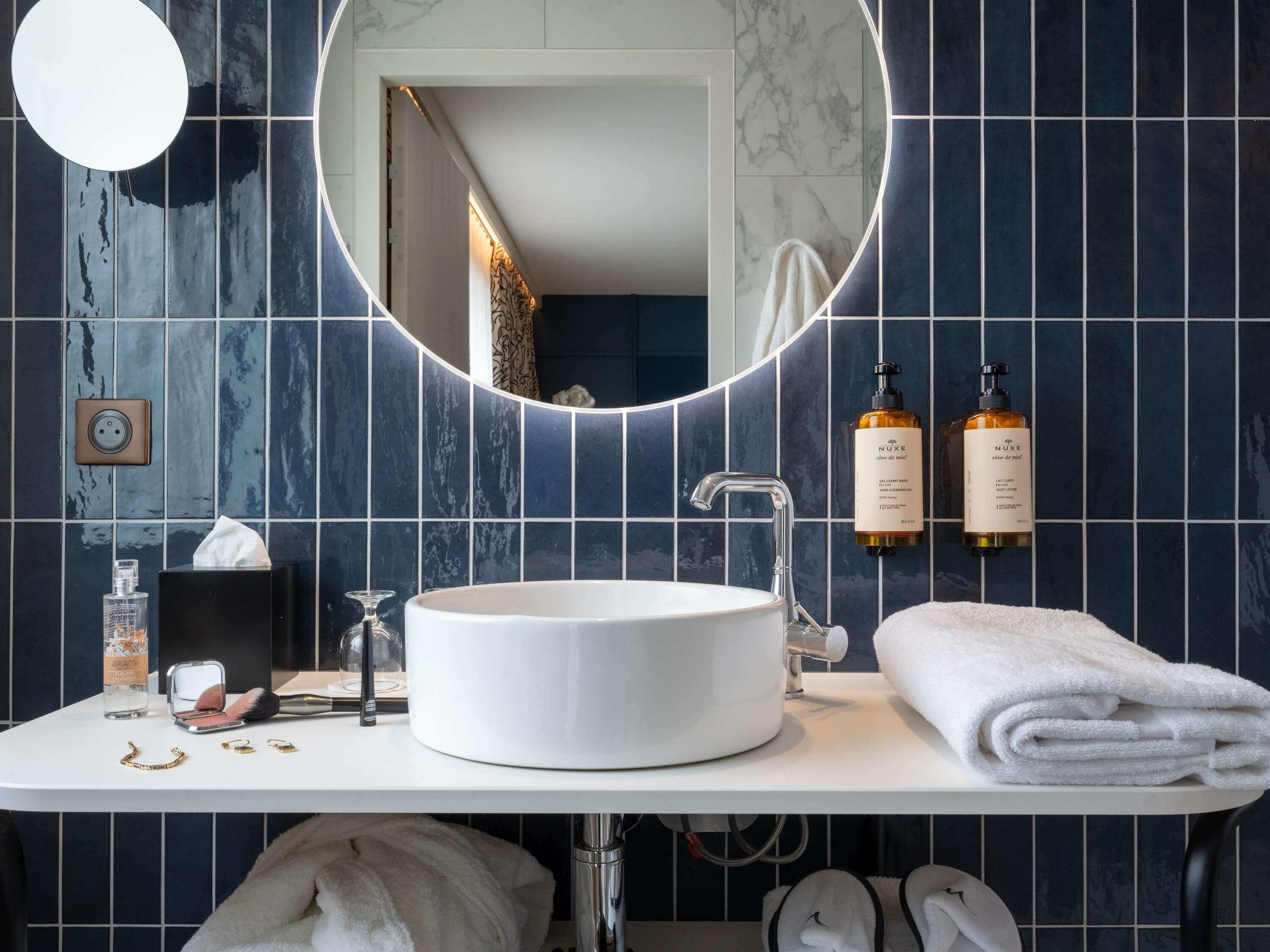 Un baño con un lavabo blanco redondo, un espejo con marco circular y dos botellas de crema corporal Nuxe en la pared.