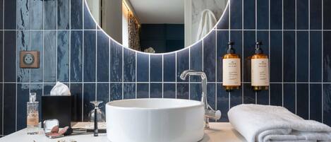 Un baño con un lavabo blanco redondo, un espejo con marco circular y dos botellas de crema corporal Nuxe en la pared.