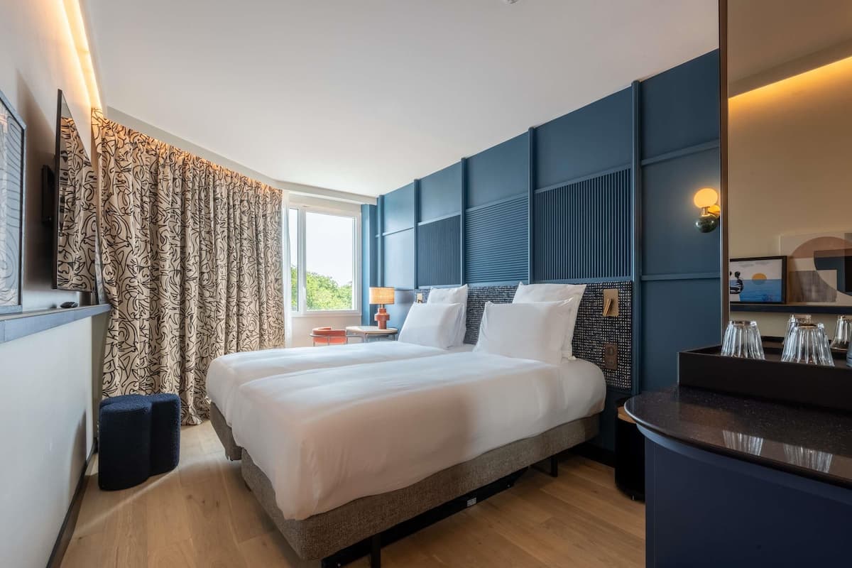 Une chambre d’hôtel avec un grand lit, une table de chevet, une lampe, un miroir et une fenêtre avec des rideaux.