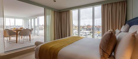 Habitación de hotel con una cama grande, zona de comedor con mesa y sillas, y vistas de la ciudad a través de amplios ventanales.