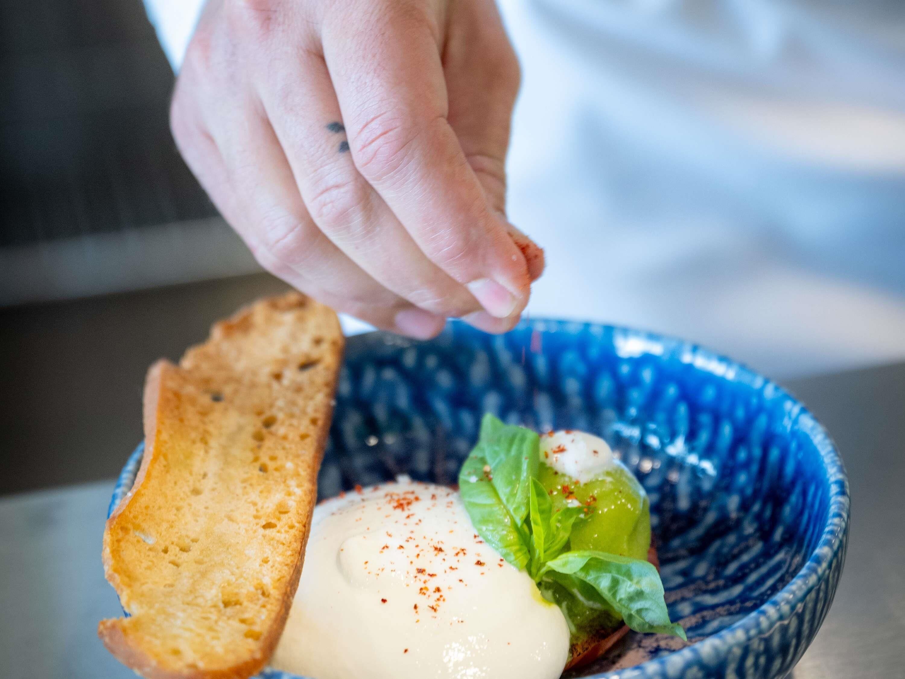 A hand sprinkling seasoning on a dish containing a slice of bread, a dollop of sauce, and a green leaf.